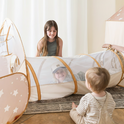 Three young children play indoors with the Tiny Land® Discovery 5-in-1 Play Tunnel With no Balls. One girl smiles at the end of the tunnel, while two younger kids, one inside and one outside, look at her.