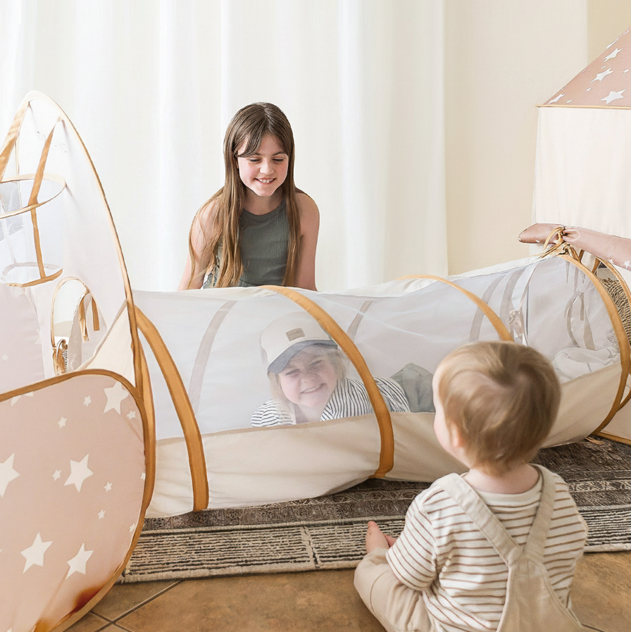 Three young children play indoors with the Tiny Land® Discovery 5-in-1 Play Tunnel With no Balls. One girl smiles at the end of the tunnel, while two younger kids, one inside and one outside, look at her.