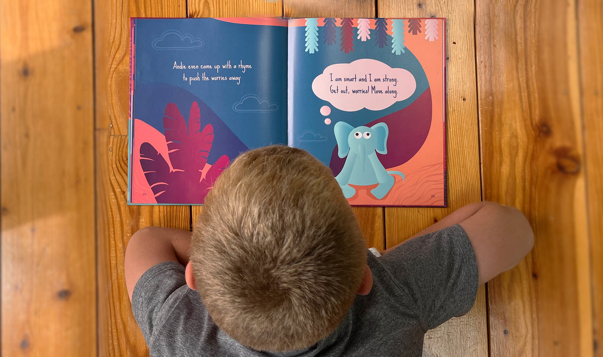A child reads the Andie & the Worries – Picture Book, which shows a blue elephant in a speech bubble saying, I am smart and I am strong. Get out, worries! Move along.