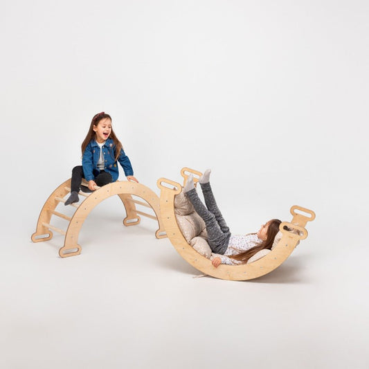 Two young girls engage in imaginative play; one smiles atop the beige Climbing Arch & Rocker Balance - Montessori Climbers for Kids 1-7 y.o., while the other reclines with her legs up on a curved balance board. The background is plain and light-colored.
