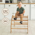 A young boy plays on a Climbing Arch & Rocker Balance - Montessori Climbers for Kids 1-7 y.o. – Beige in a bright kitchen with white cabinets and light wood countertop. Two circular badges nearby read CPC Certified and EU surrounded by stars.