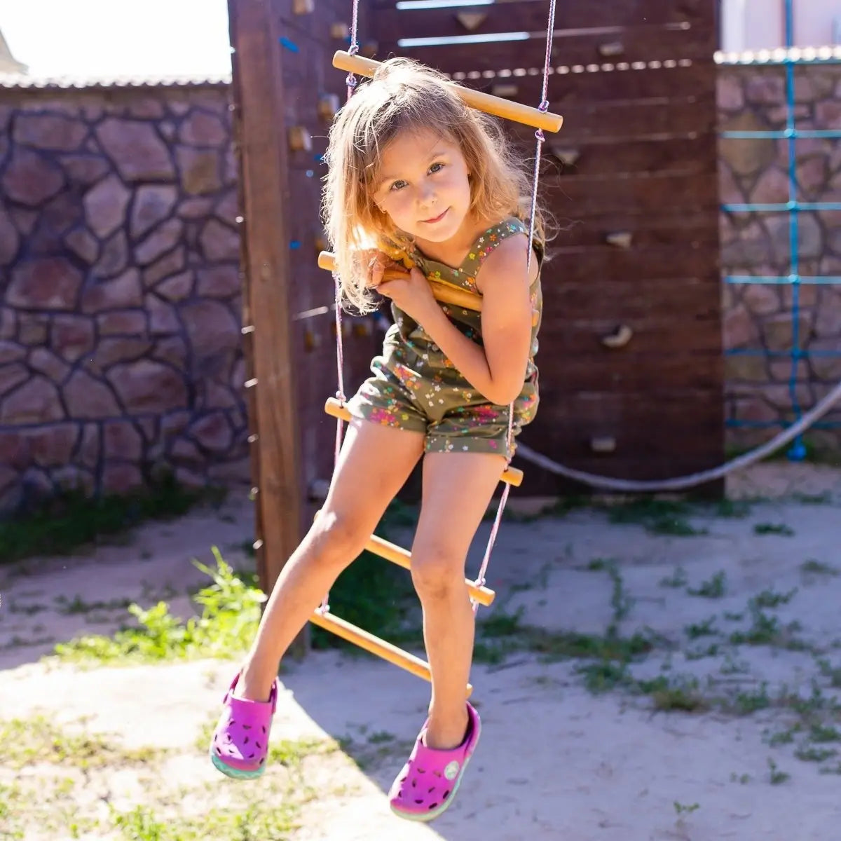 A young girl in a green outfit and pink shoes sits on the Climbing rope ladder for kids 3-9 y.o. at a playground, smiling gently with sunlight on her face—enjoying an activity that promotes muscle strengthening and supports child development.