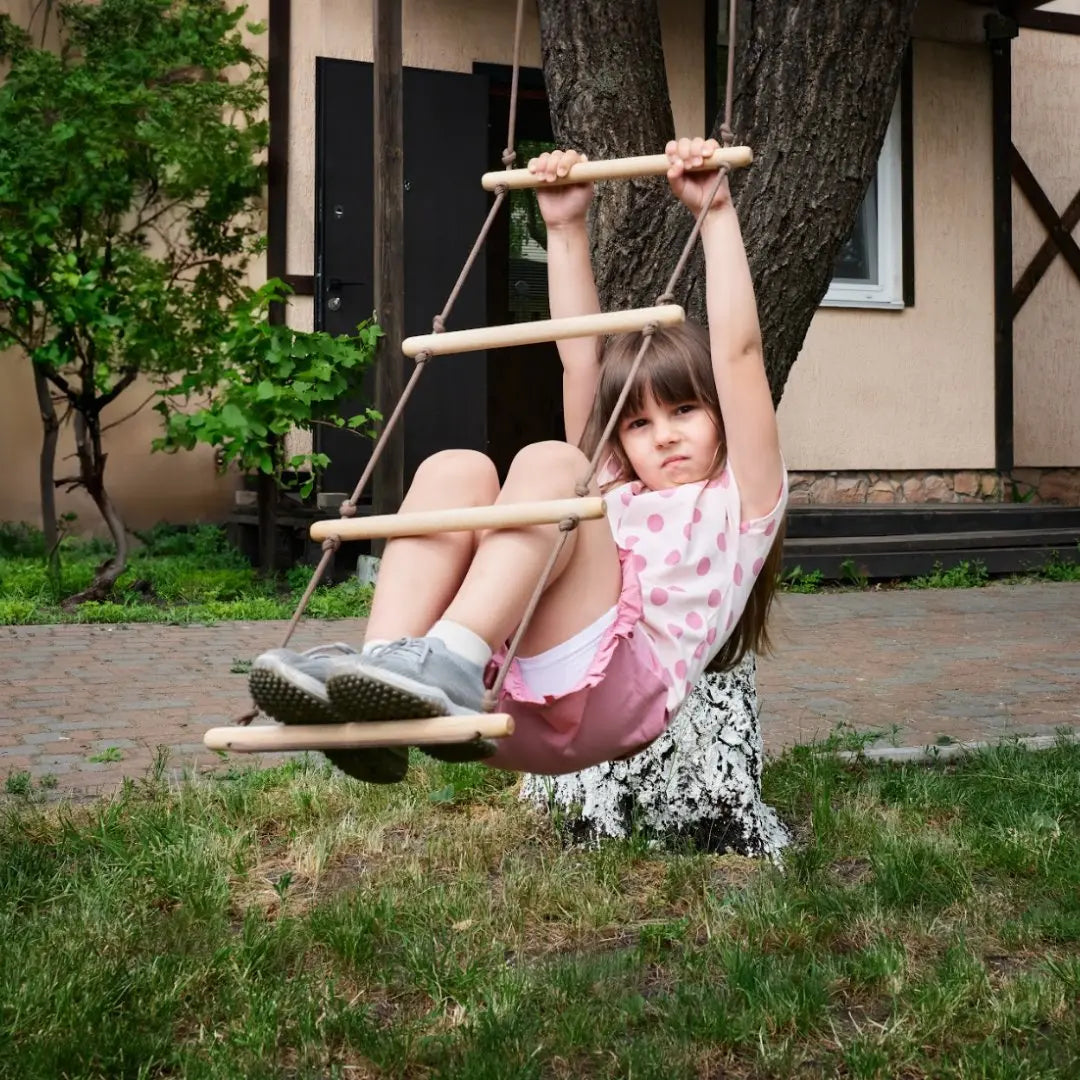 A young girl in a pink outfit hangs from a Climbing rope ladder for kids 3-9 y.o., which is attached to a tree in the yard, looking at the camera with a serious expression—a playful moment that also supports child development and muscle strengthening.