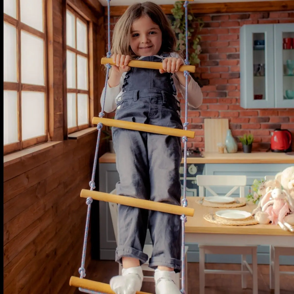 A smiling child in overalls climbs a Climbing rope ladder for kids 3-9 y.o. by Goodevas indoors, with a cozy kitchen featuring a brick wall, wooden table, and plush toys in the background. Sunlight streams through large windows on the left, creating a warm environment perfect for child development and muscle strengthening.