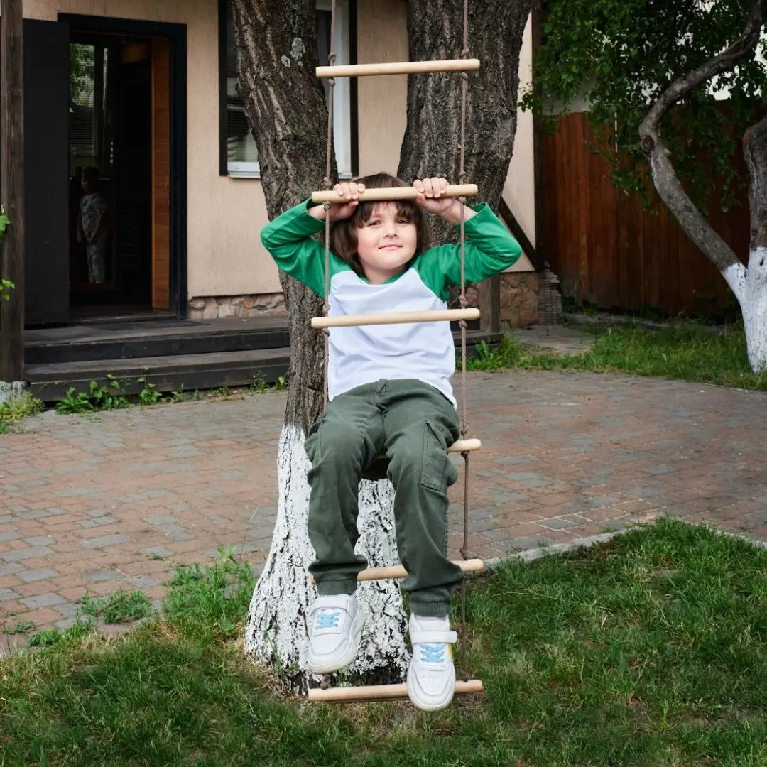 A young boy in green pants and a white-green shirt climbs the Climbing rope ladder for kids 3-9 y.o., attached to a tree in a backyard with grass, pavement, and a wooden house in the background, promoting muscle strengthening and supporting healthy child development.
