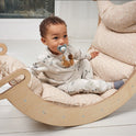 A baby with a pacifier sits comfortably in a cushioned wooden rocking chair indoors, wearing light pajamas with small patterns and nestled on the Cushion for Climbing Arch & Rocker Balance (Cushion Only).