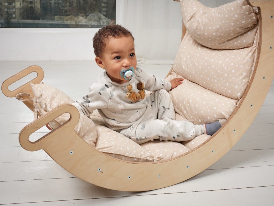 A baby with a pacifier sits comfortably in a cushioned wooden rocking chair indoors, wearing light pajamas with small patterns and nestled on the Cushion for Climbing Arch & Rocker Balance (Cushion Only).