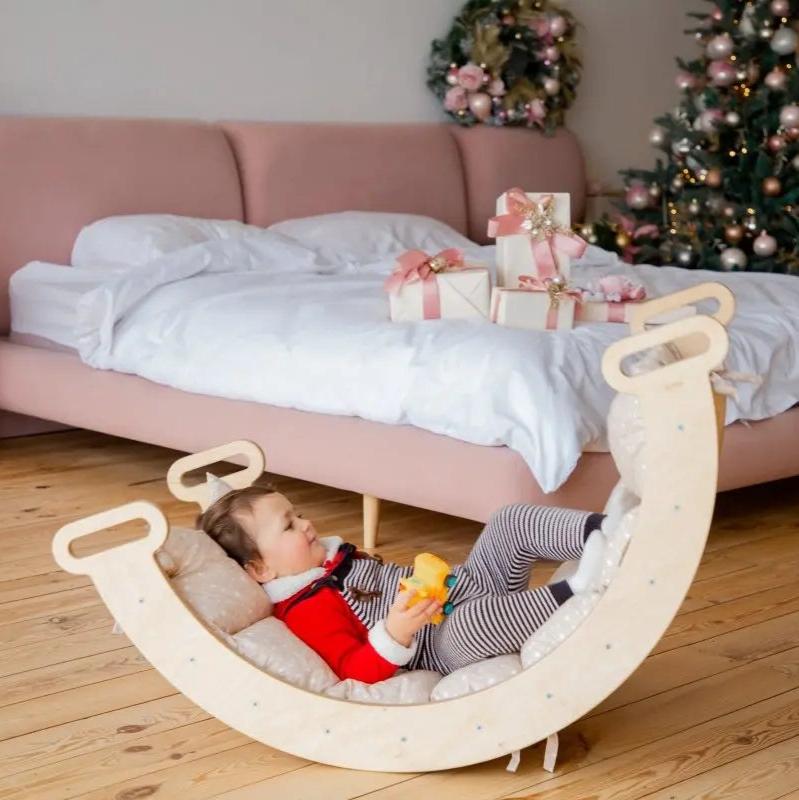 A baby in red pajamas lies in a wooden rocker fitted with the Cushion for Climbing Arch & Rocker Balance (Cushion Only), holding a toy, in a cozy bedroom with a pink sofa, wrapped gifts, and a decorated Christmas tree in the background.
