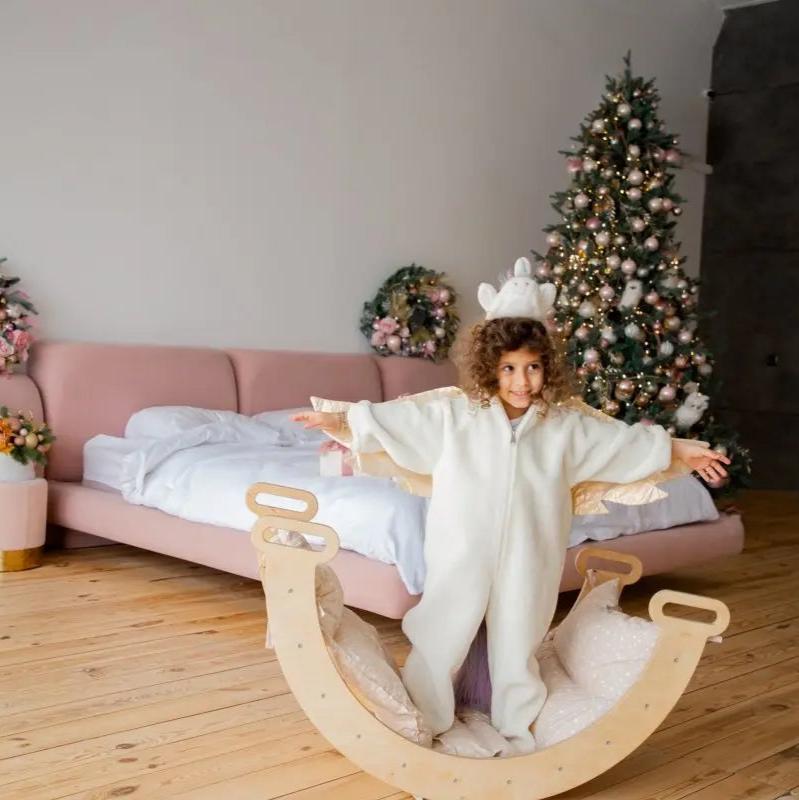 A young child in a white onesie stands on a wooden rocker with arms outstretched, balanced atop the Cushion for Climbing Arch & Rocker Balance (Cushion Only) in a warm bedroom decorated with a Christmas tree, festive wreaths, and a pink bed.