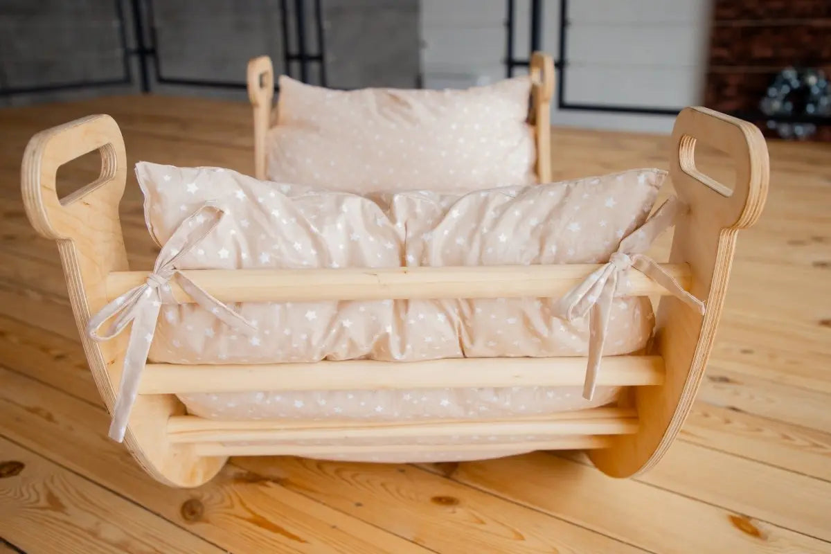 A small wooden cradle with beige polka-dot bedding and pillows tied with ribbons sits on a light wooden floor in a cozy room, perfectly complemented by our Cushion for Climbing Arch & Rocker Balance (Cushion Only) to enhance your little one’s comfort.