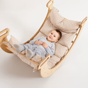A smiling baby in grey pajamas lies comfortably on a wooden rocking chair, cozily supported by the Cushion for Climbing Arch & Rocker Balance (Cushion Only), featuring beige star-patterned pillows and a soft Montessori cushion against a white background.
