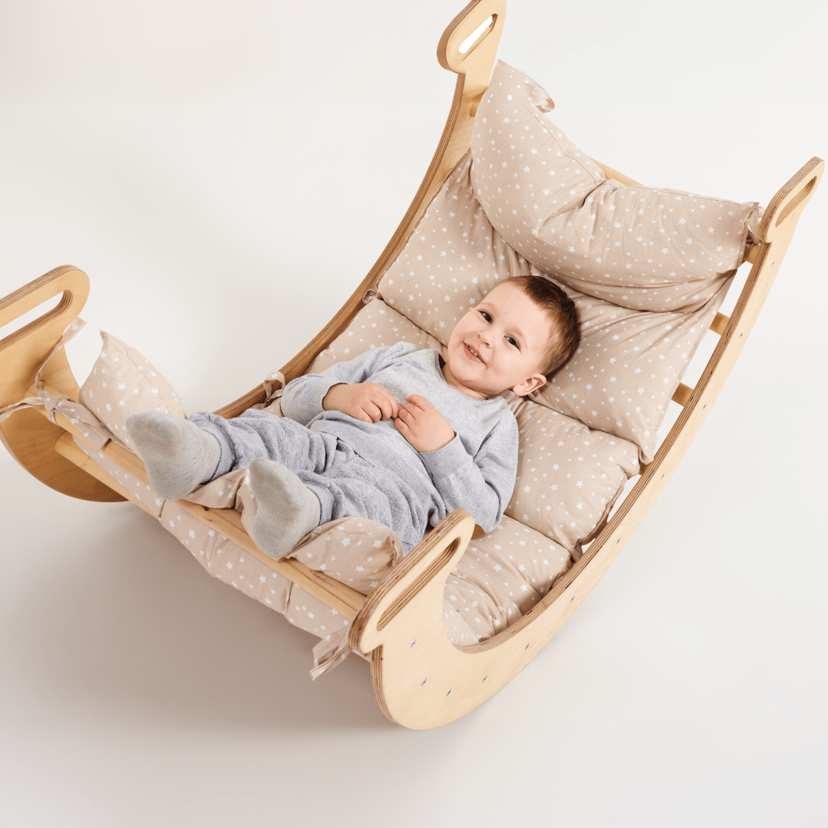 A smiling baby in grey pajamas lies comfortably on a wooden rocking chair, cozily supported by the Cushion for Climbing Arch & Rocker Balance (Cushion Only), featuring beige star-patterned pillows and a soft Montessori cushion against a white background.