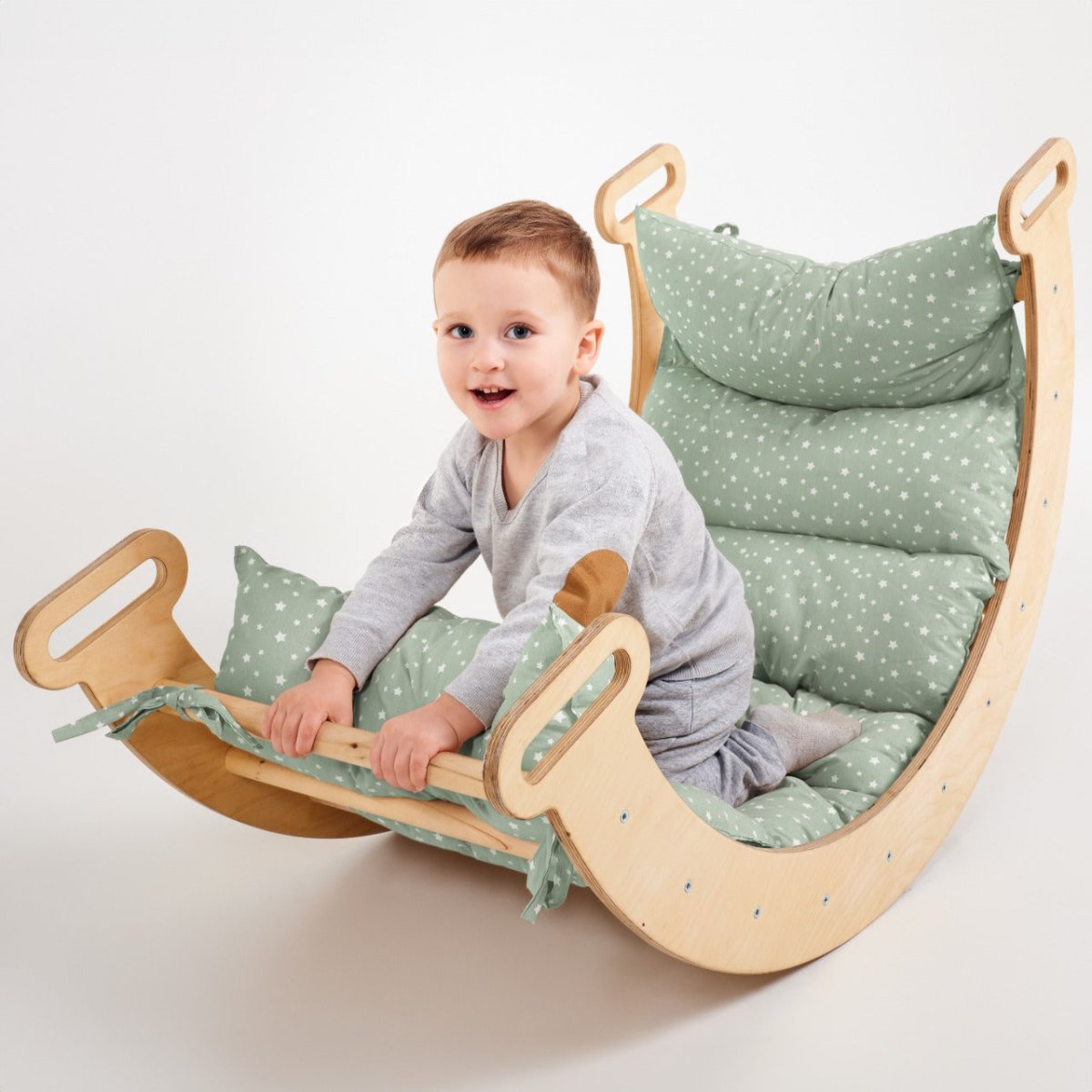 A young child in gray pajamas sits on a wooden rocking chair with a green Cushion for Climbing Arch & Rocker Balance (Cushion Only), smiling and holding onto the edge.