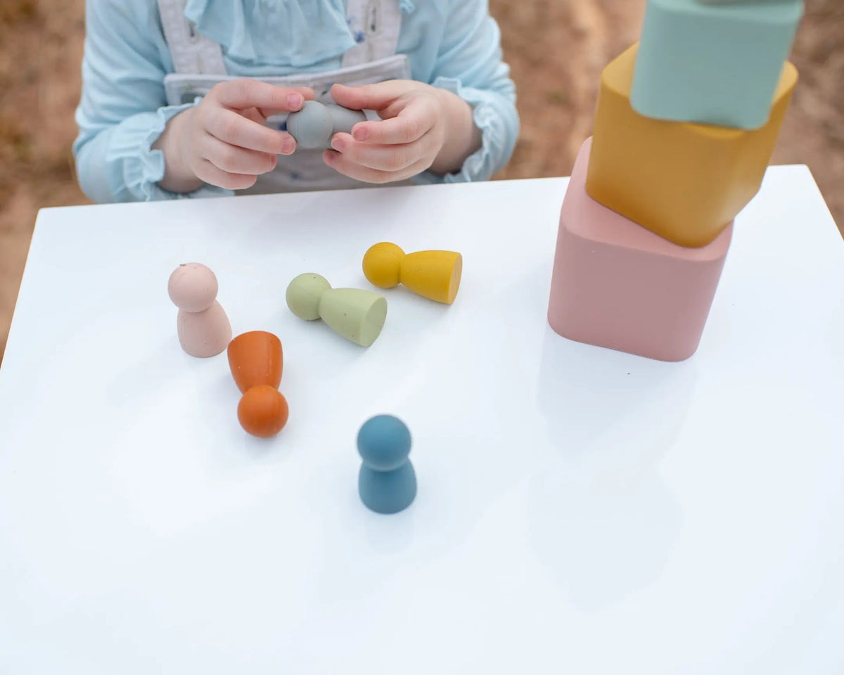 A child holds a small grey Silicone Mini Peg Dolls piece near a white table scattered with five colorful silicone peg dolls and a stack of three square silicone blocks.