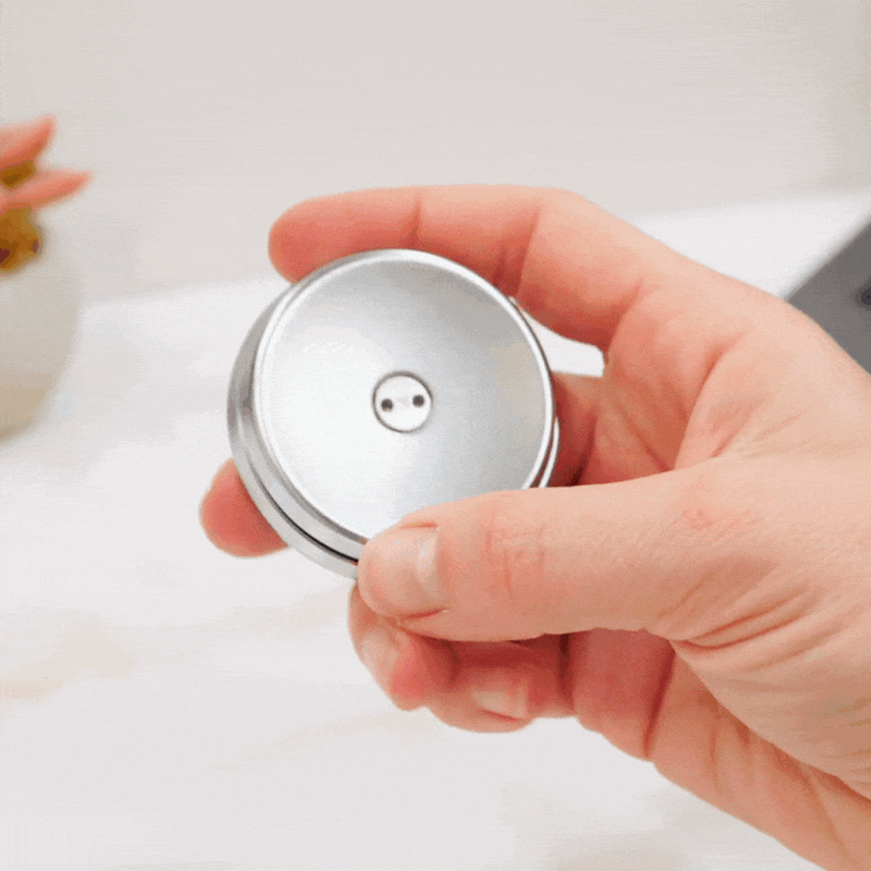 A hand spins the ONO Slider Steel, a sleek minimalist silver fidget toy, against a white background. In the blurred background are a plant, part of a laptop, and small magnetized disks from another fidget toy.