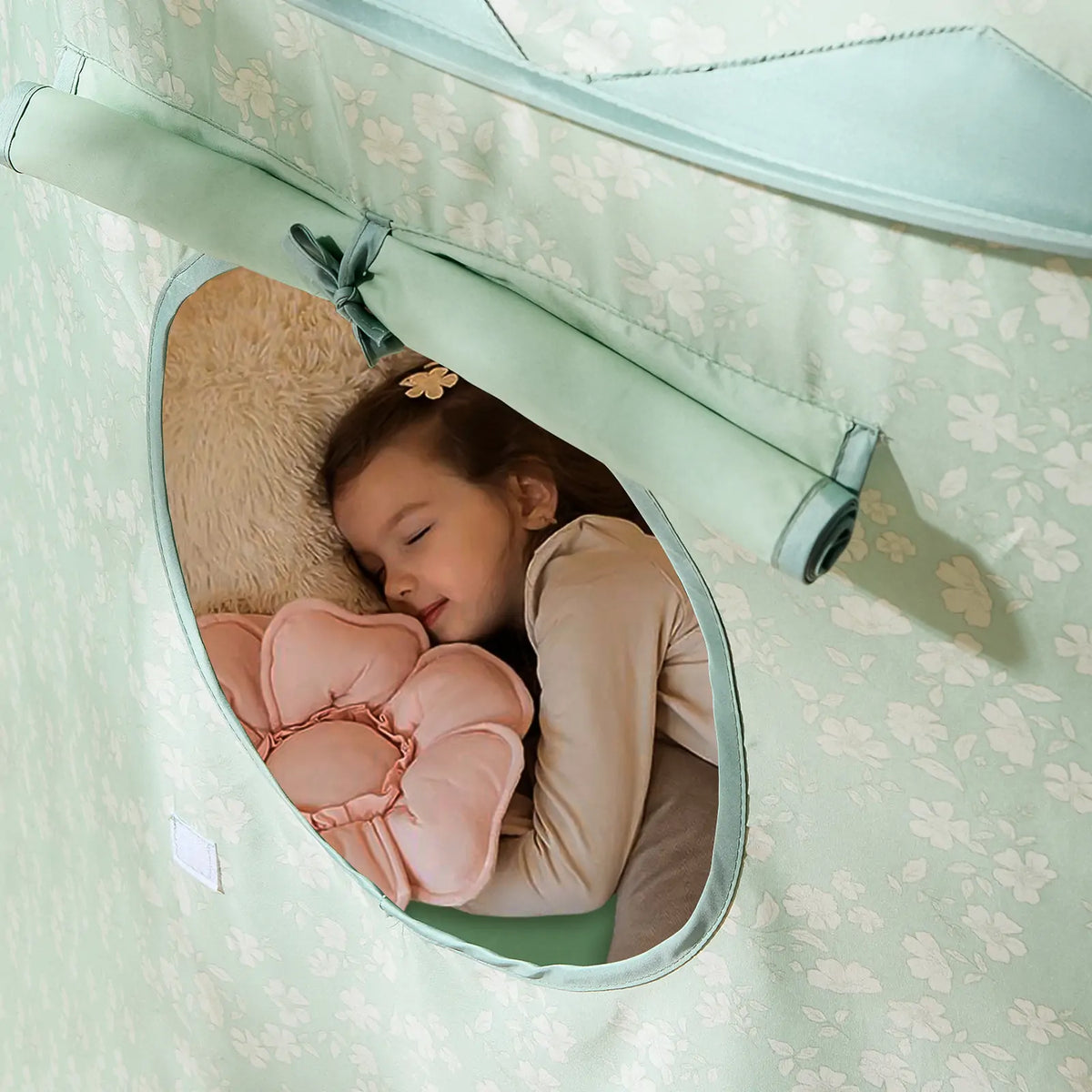 A young child sleeping inside a mint green floral Starry Indoor Play Tent for Toddlers, resting their head on a pink flower-shaped pillow.