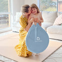 A woman in a yellow polka-dot dress kisses a smiling toddler holding the blue circular carrying case for their Starry Indoor Play Tent for Toddlers.
