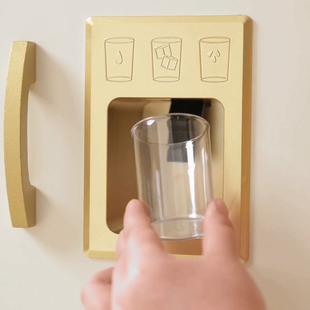 A hand wearing a chef hat holds a plastic cup under the dispenser of a PopJoy Play Kitchen with Lights & Sounds refrigerator featuring icons for water, ice, and crushed ice.