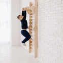 A young boy in dark clothes hangs from a wooden climbing ladder with the Gym Addition to the Climbing Ladders, which is attached to a white brick wall in a bright room with a white floor.