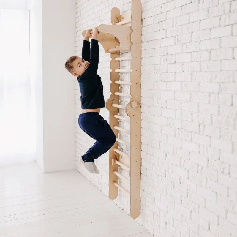 A young boy in dark clothes hangs from a wooden climbing ladder with the Gym Addition to the Climbing Ladders, which is attached to a white brick wall in a bright room with a white floor.