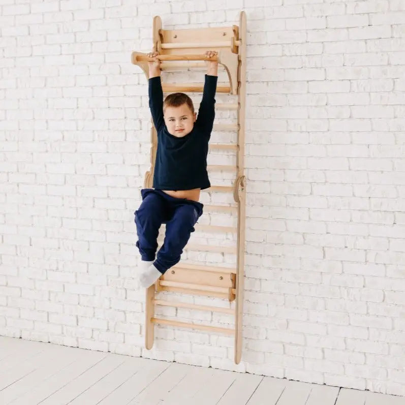 A young boy in a dark outfit hangs by his hands from wooden wall bars, which are equipped with the Gym Addition to the Climbing Ladders, attached to a white brick wall, with his feet off the ground—enjoying some indoor entertainment.