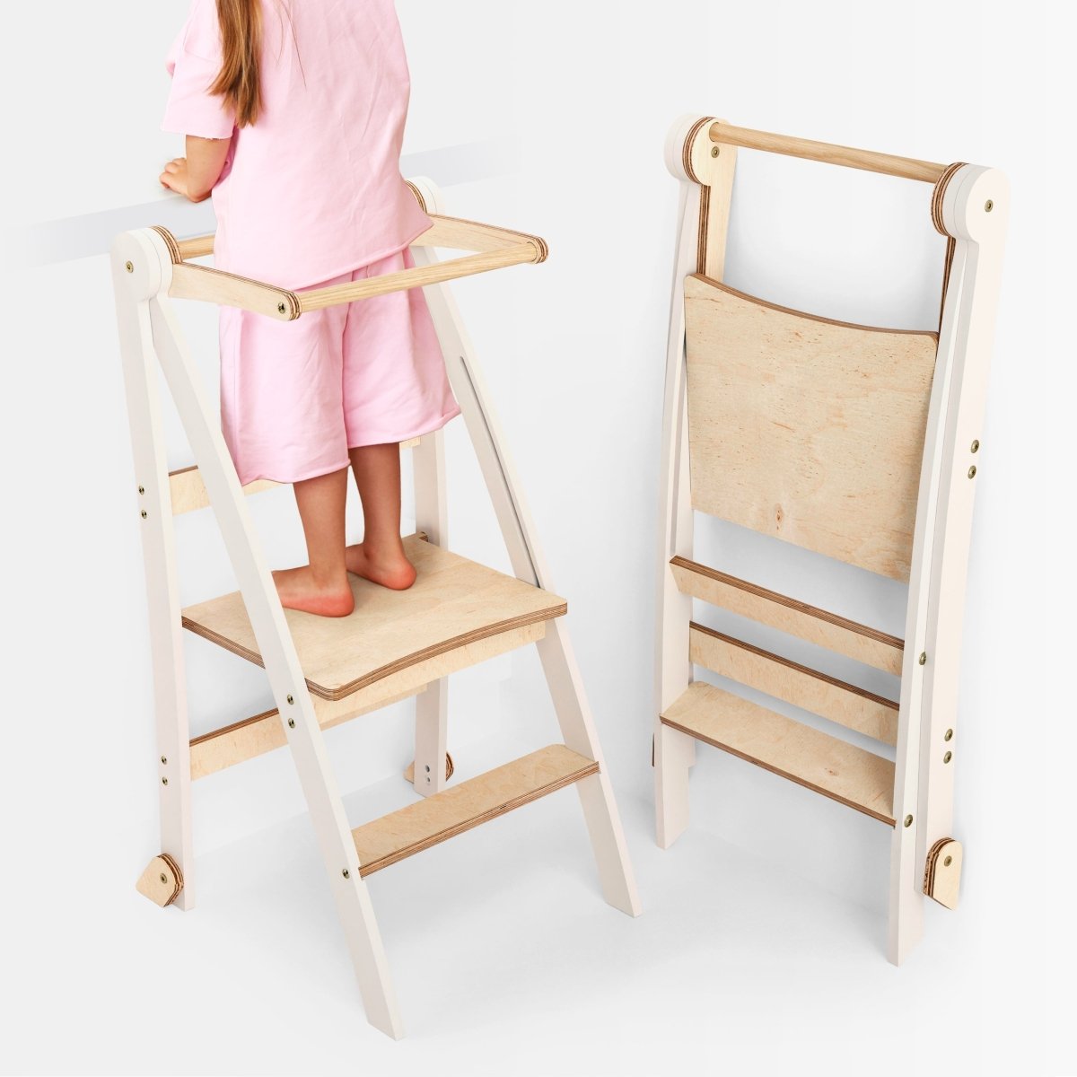 A young child stands on the Helper Tower: A Multi-functional Foldable Step Stool for Kids, featuring white coloring with natural wood accents and safety railings; beside it, the same stool is shown folded flat for easy storage.