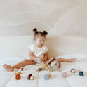 A toddler sits on a bed playing with a Silicone Rainbow Stacking Toy (10 pc) and several small, mushroom-shaped wooden pieces.