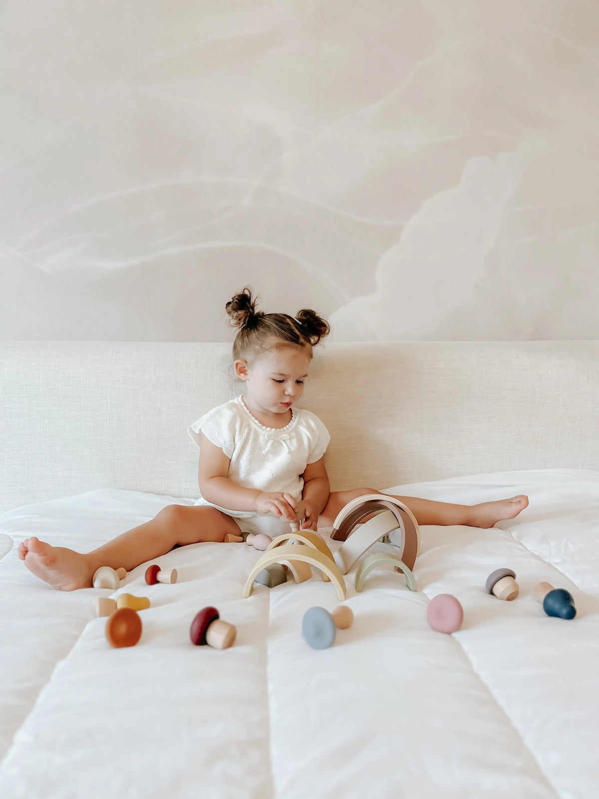 A toddler sits on a bed playing with a Silicone Rainbow Stacking Toy (10 pc) and several small, mushroom-shaped wooden pieces.