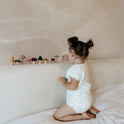 A toddler kneels on a bed, placing small wooden Mushroom Mix & Match Sorting Toy (24 pc) mushrooms in a row along the headboard to practice color sorting.
