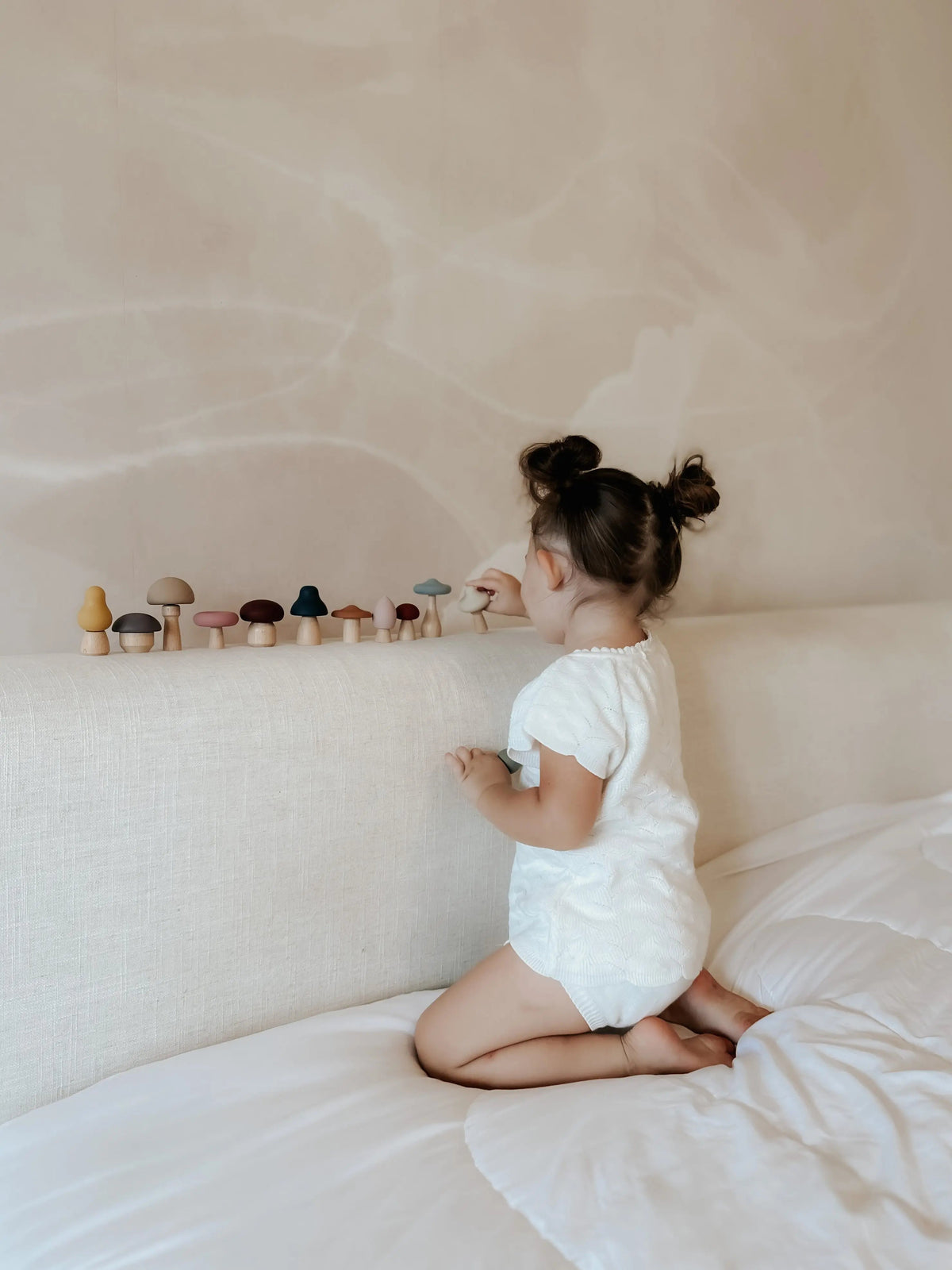 A toddler kneels on a bed, placing small wooden Mushroom Mix & Match Sorting Toy (24 pc) mushrooms in a row along the headboard to practice color sorting.