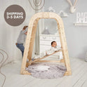 Two young children play on the Indoor Wooden Playground for Children – 6in1 Playground + Swings Set + Slide Board inside a softly decorated room. One child stands while the other lies on the suspended slide board, smiling. The space features gentle, neutral tones and a round pillow on the floor.