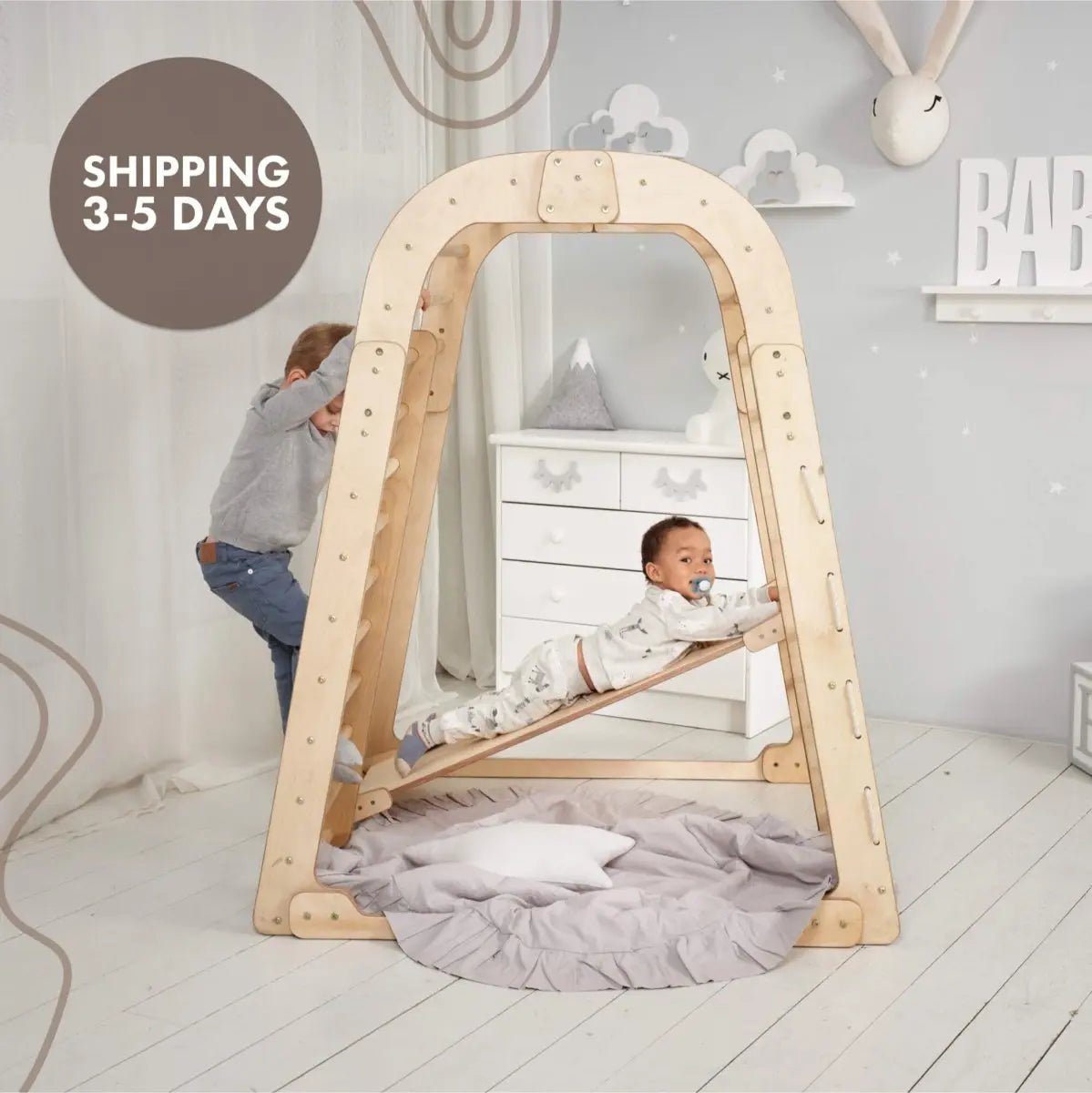 Two young children play on the Indoor Wooden Playground for Children – 6in1 Playground + Swings Set + Slide Board inside a softly decorated room. One child stands while the other lies on the suspended slide board, smiling. The space features gentle, neutral tones and a round pillow on the floor.