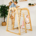 Two children play on the Indoor Wooden Playground for Children – 6in1 Playground + Swings Set + Slide Board; one climbs the ladder while the other navigates the rope net. This versatile indoor playset, designed to be a mini kids’ playground, is set in a cozy room featuring a basketed plant and shelves filled with books in the background.