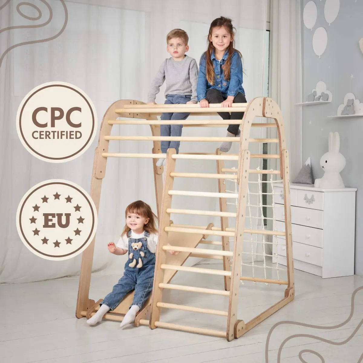 Three children play on the Indoor Wooden Playground for Children - 6in1 Playground + Swings Set + Slide Board in a bright, airy room decorated with toys. Two kids are at the top of the wooden Montessori-inspired playset, while one climbs underneath. Text in the image displays “CPC Certified” and “EU,” each highlighted in star-shaped round badges.