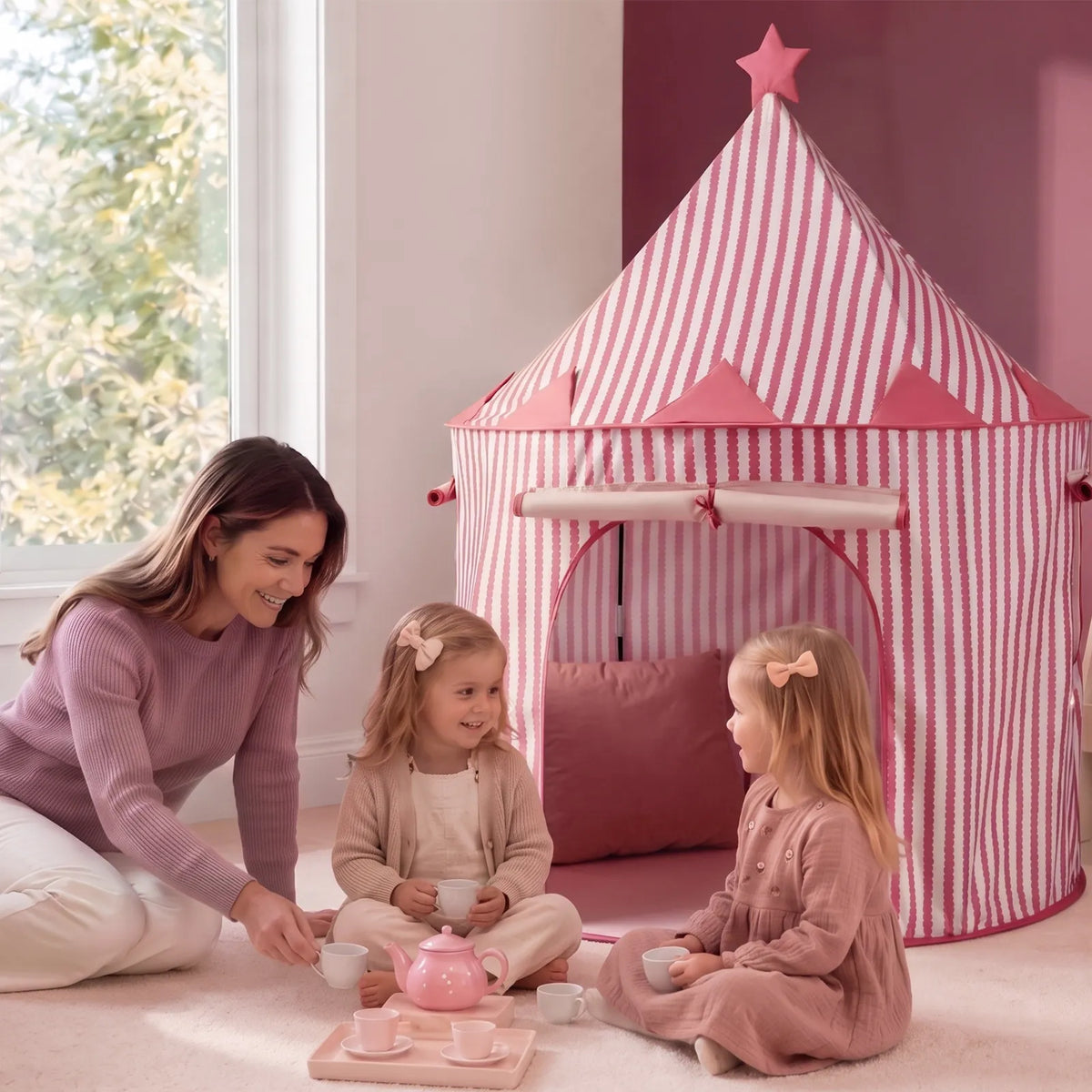 A woman and two young girls sit on the floor having a tea party next to a pink and white striped Starry Indoor Play Tent for Toddlers.