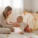 A young child crawls out of a 68″ Adventure Crawl Tunnel toward a smiling woman sitting on the floor.