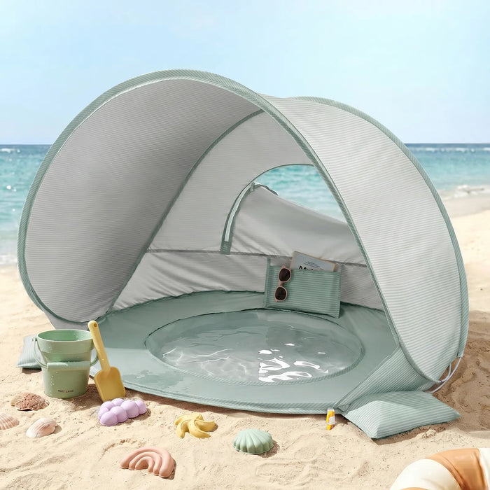 Green 3-in-1 Baby Beach Tent with Splash Pool sitting on a sandy beach with plastic sand toys in the foreground.