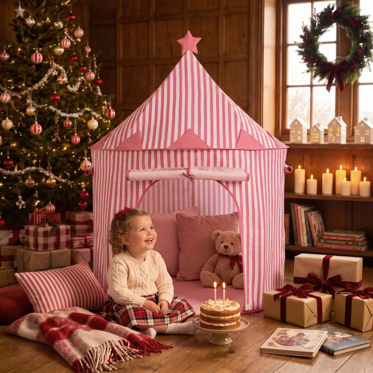 A toddler sits on the floor next to a pink and white striped Starry Indoor Play Tent for Toddlers, with a birthday cake and a teddy bear inside.