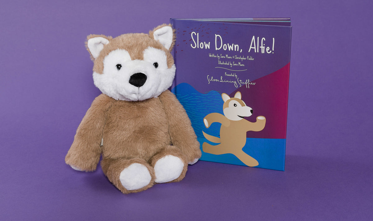 A brown and white wolf plush sitting next to a children’s book titled Slow Down, Alfie!, part of the Alfie the Wolf Plush + Book set.
