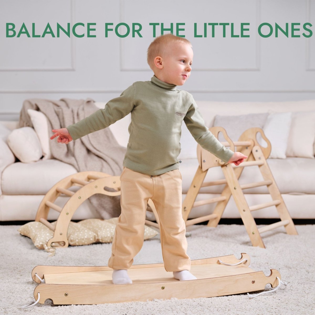 A toddler in beige pants and a green top balances on the 4in1 Montessori Climbing Set: Pikler Triangle, Arch, Slide Board, and Cushion for Toddlers (1–3 Years), with a cozy sofa in the background. Text above reads, BALANCE FOR THE LITTLE ONES.