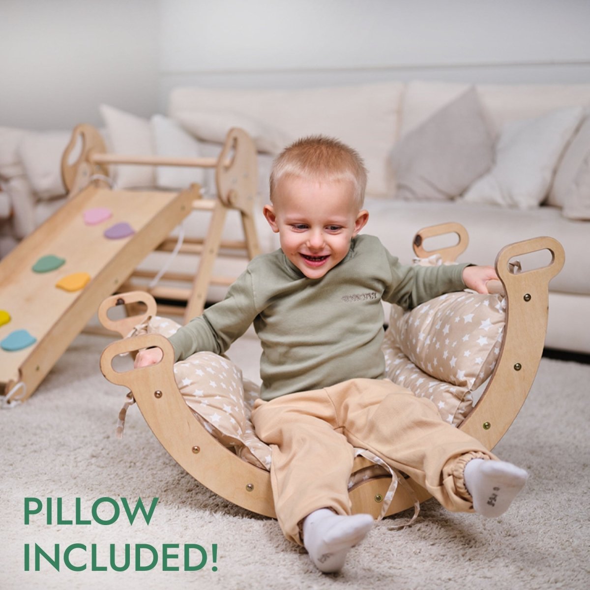 A smiling toddler sits in the 4in1 Montessori Climbing Set: Pikler Triangle, Arch, Slide Board, and Cushion for Toddlers (1–3 Years) in a cozy living room. Colorful grips are visible. Text reads PILLOW INCLUDED!.
