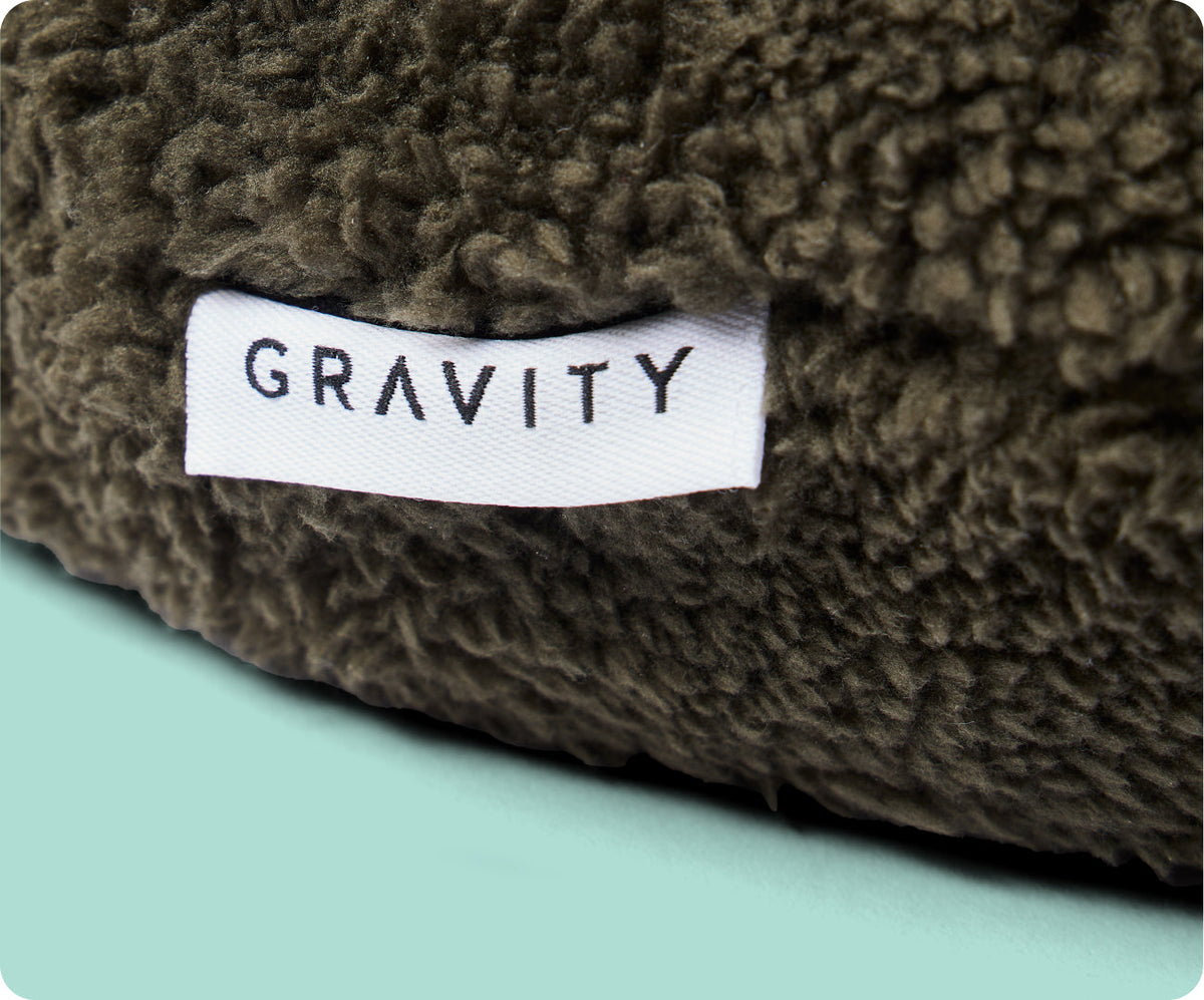Close-up of a textured dark olive fabric with a white label reading GRAVITY stitched onto the corner of the Gravity Weighted Neck Pillow (4 lb).