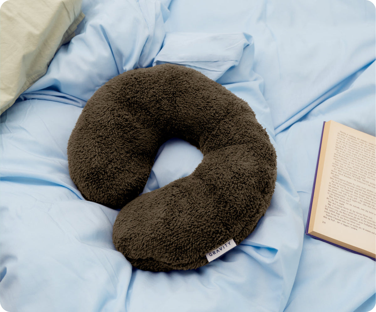 Gravity Weighted Neck Pillow (4 lb) in a U-shaped brown fuzzy design resting on light blue bedding next to an open book.