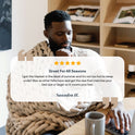 A man sits at a table with a laptop, wrapped in a Knit Weighted Blanket. A five-star review praises its comfort in every season and suggests choosing the right size. A coffee mug and plant complete the cozy scene.