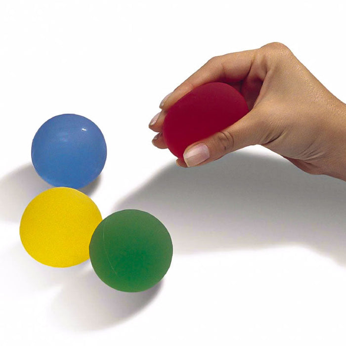 A hand squeezes a red THERABAND Hand Exerciser Grip Ball to improve grip strength, with blue, yellow, and green balls resting on a white surface nearby.