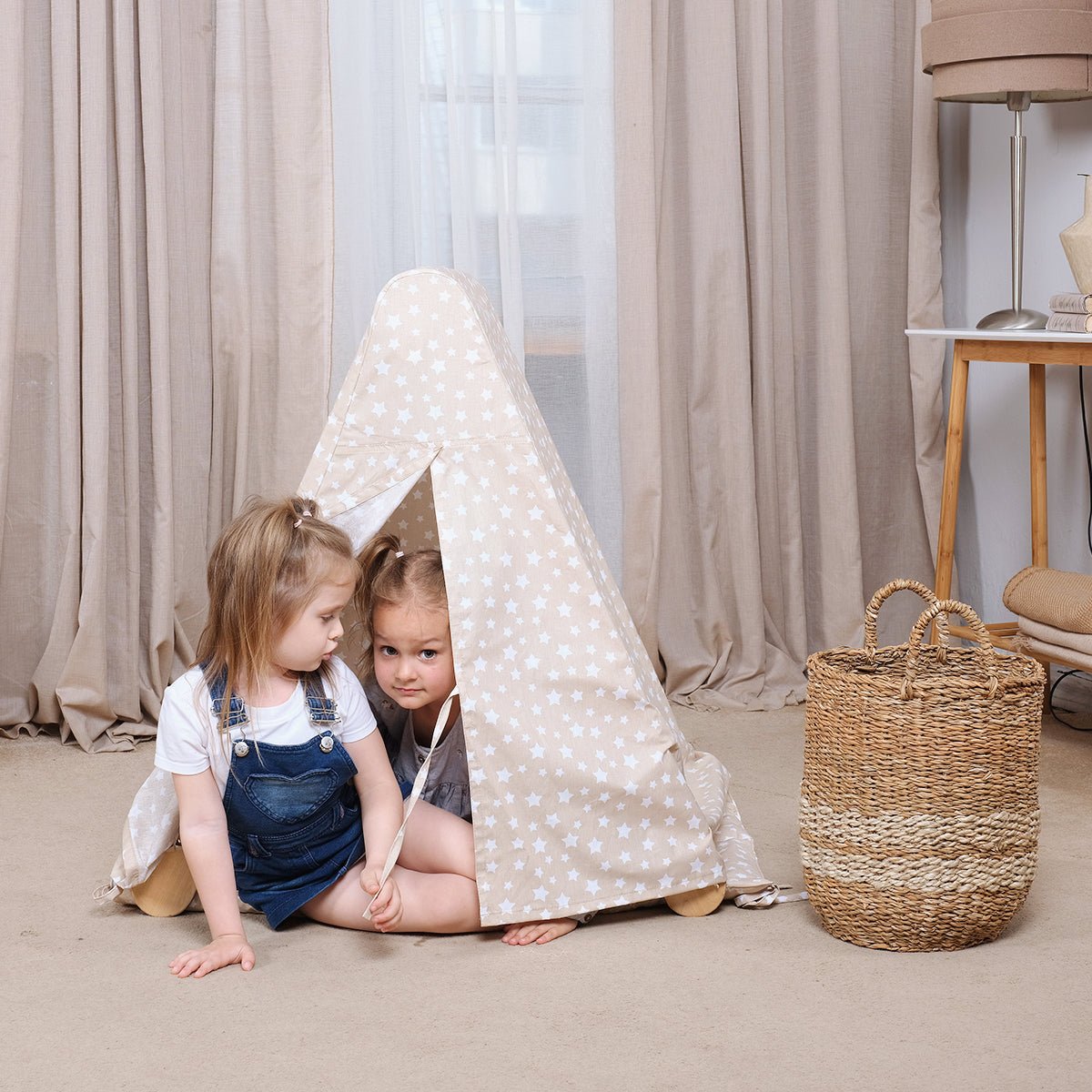Two young children sit inside a Play Tent Cover with Mat for Climbing Triangle Ladder, decorated with white stars. One child in overalls sits just outside the tent, while the other peeks out from within. A woven basket rests on the floor beside them, next to a Montessori triangle ladder. Beige curtains hang in the background.