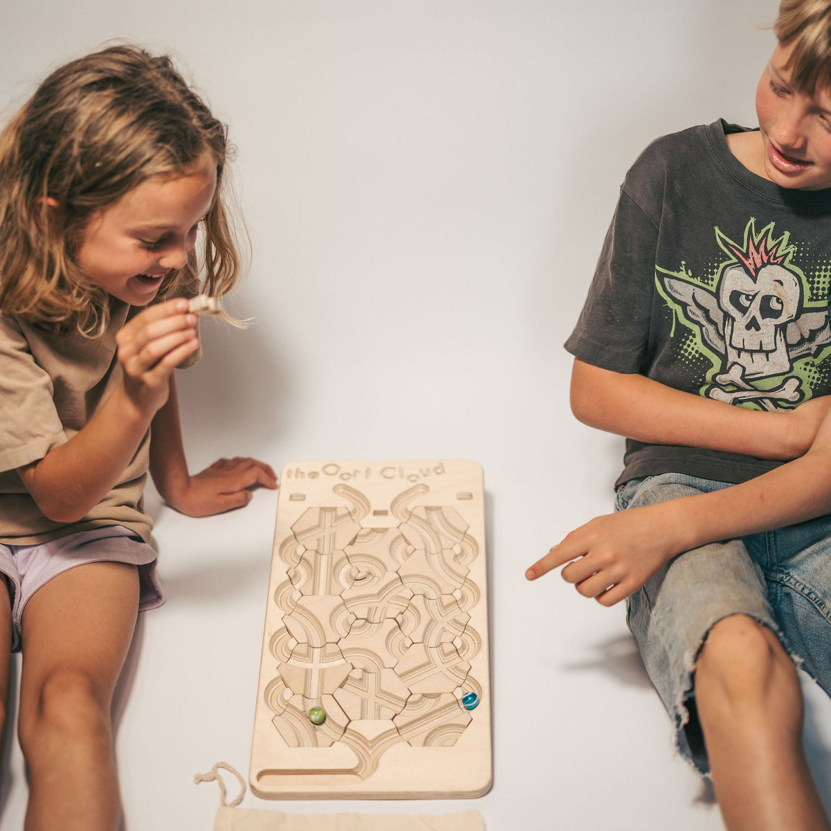 Two children sit on the floor playing with The Oort Cloud – Wooden Marble Run Puzzle, a handmade wooden marble run featuring circular tracks and colorful marbles.