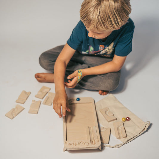 A child sits on the floor building an Oortle – Mini Wooden Marble Run Puzzle, carefully assembling the brain teaser with various tracks and marbles.