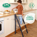A young child stands on the Toddler Tower: Foldable Step Stool for Toddlers - Gray in a kitchen, reaching for a jar on the counter. Labels indicate CPC and CE certifications and highlight the product’s organic material and cover.