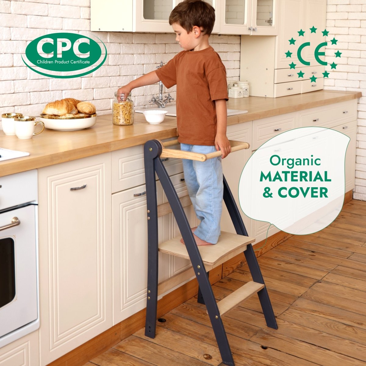 A young child stands on the Toddler Tower: Foldable Step Stool for Toddlers - Gray in a kitchen, reaching for a jar on the counter. Labels indicate CPC and CE certifications and highlight the product’s organic material and cover.